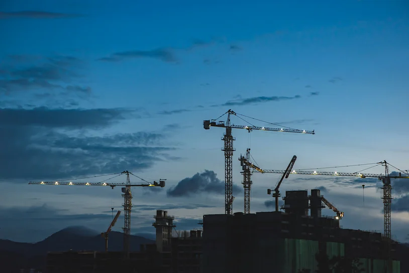 Construction cranes silhouetted against a dusk sky over a city building site
