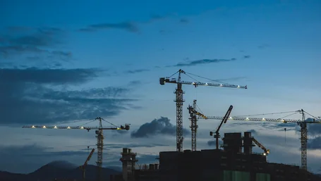 Construction cranes silhouetted against a dusk sky over a city building site