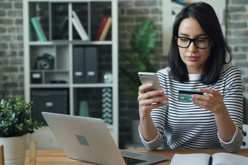 Woman in a home office holding a credit card while looking at her phone near a laptop