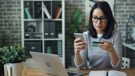 Woman in a home office holding a credit card while looking at her phone near a laptop