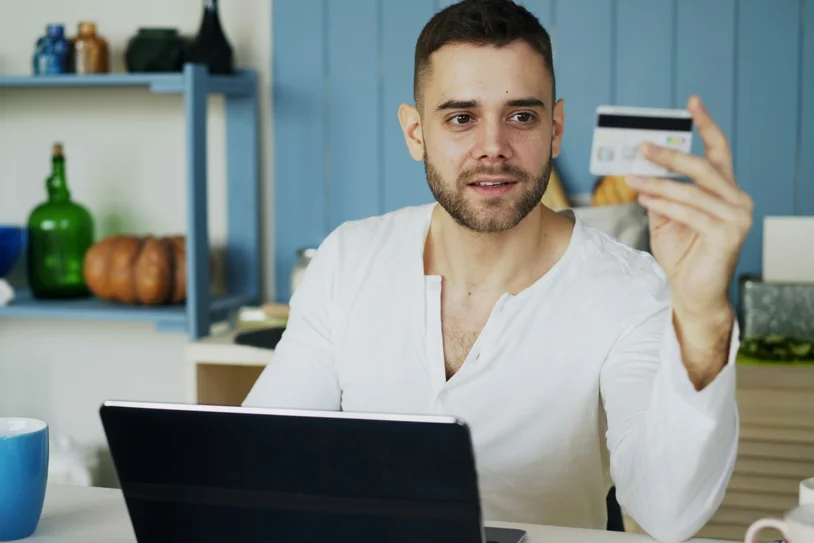 Man at a kitchen table holding a credit card while smiling at the camera near a laptop