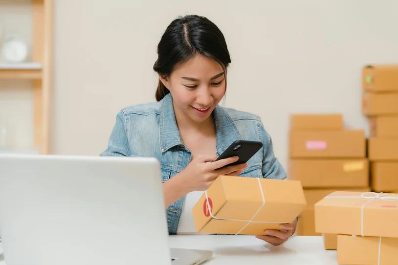 Woman checking her phone while holding a shipping box surrounded by packages