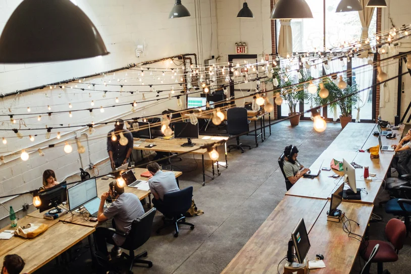 Open coworking space with people working at long shared desks under string lights