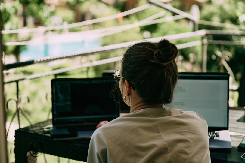 Person working on dual laptops at an outdoor desk surrounded by greenery