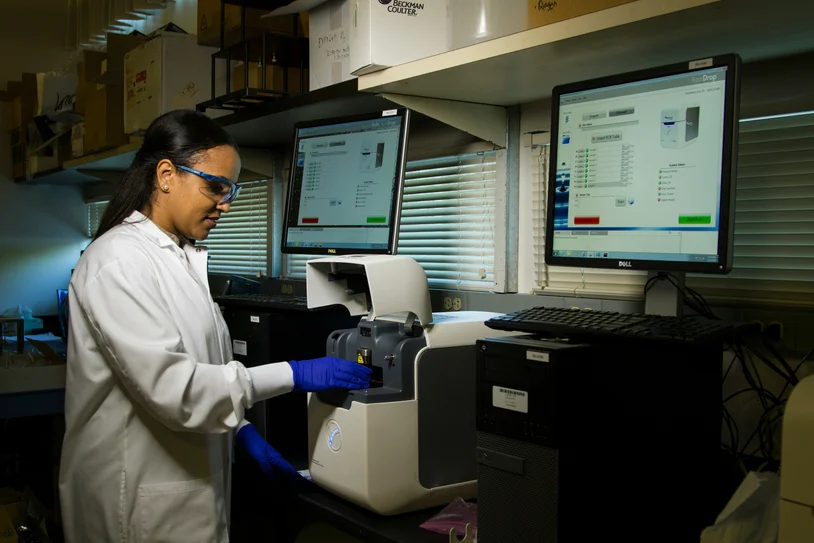 Lab technician in a white coat operating diagnostic equipment beside dual monitors