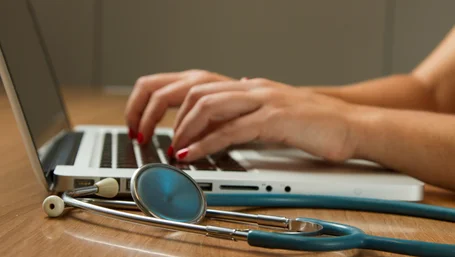 Close-up of hands typing on a laptop next to a stethoscope on a wooden desk
