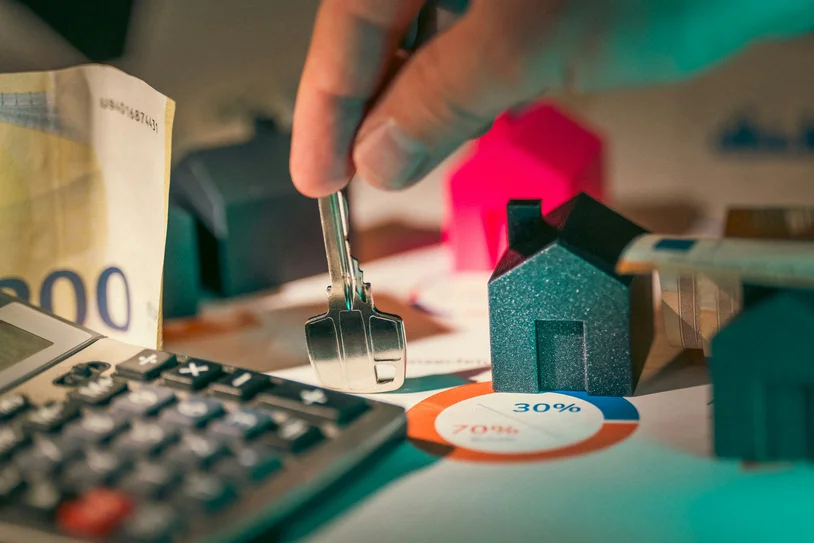Hand holding a house key over a desk with cash, a calculator, and a pie chart