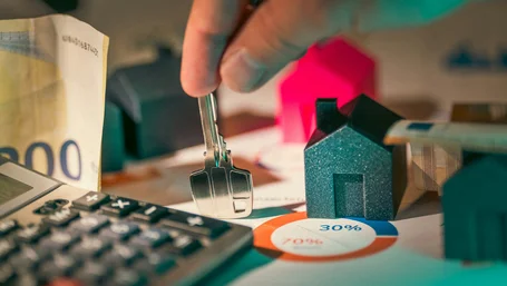 Hand holding a house key over a desk with cash, a calculator, and a pie chart