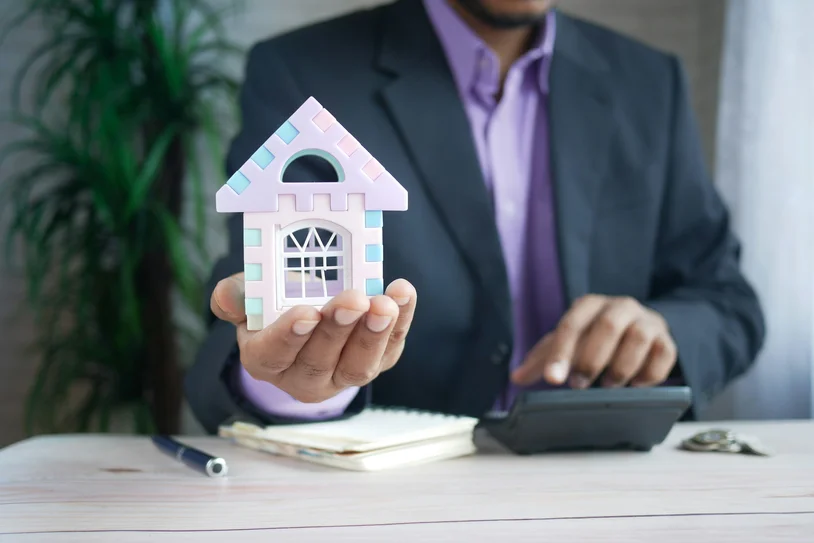 Agent in a suit holding a small house model while using a calculator at a desk
