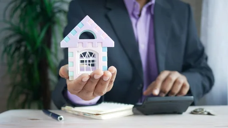 Agent in a suit holding a small house model while using a calculator at a desk